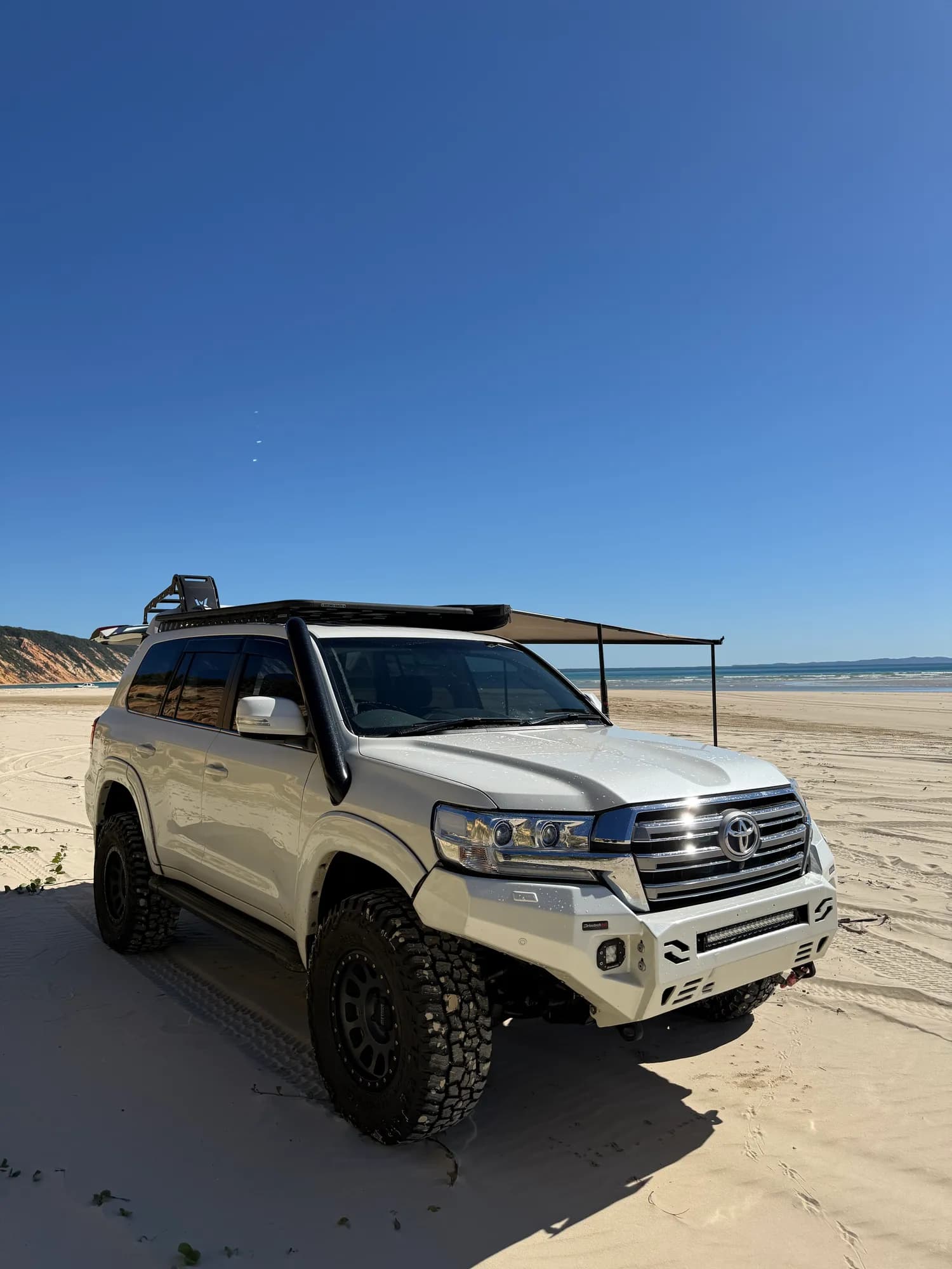 A white 200 Series with a snorkel, roof rack and awning parked on a wide beach under a clear blue sky