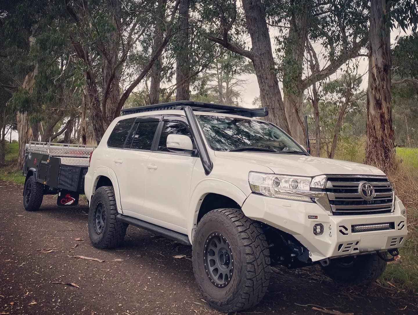 A white 200 Series towing an off-road camper trailer along a gravel track through tall gum trees