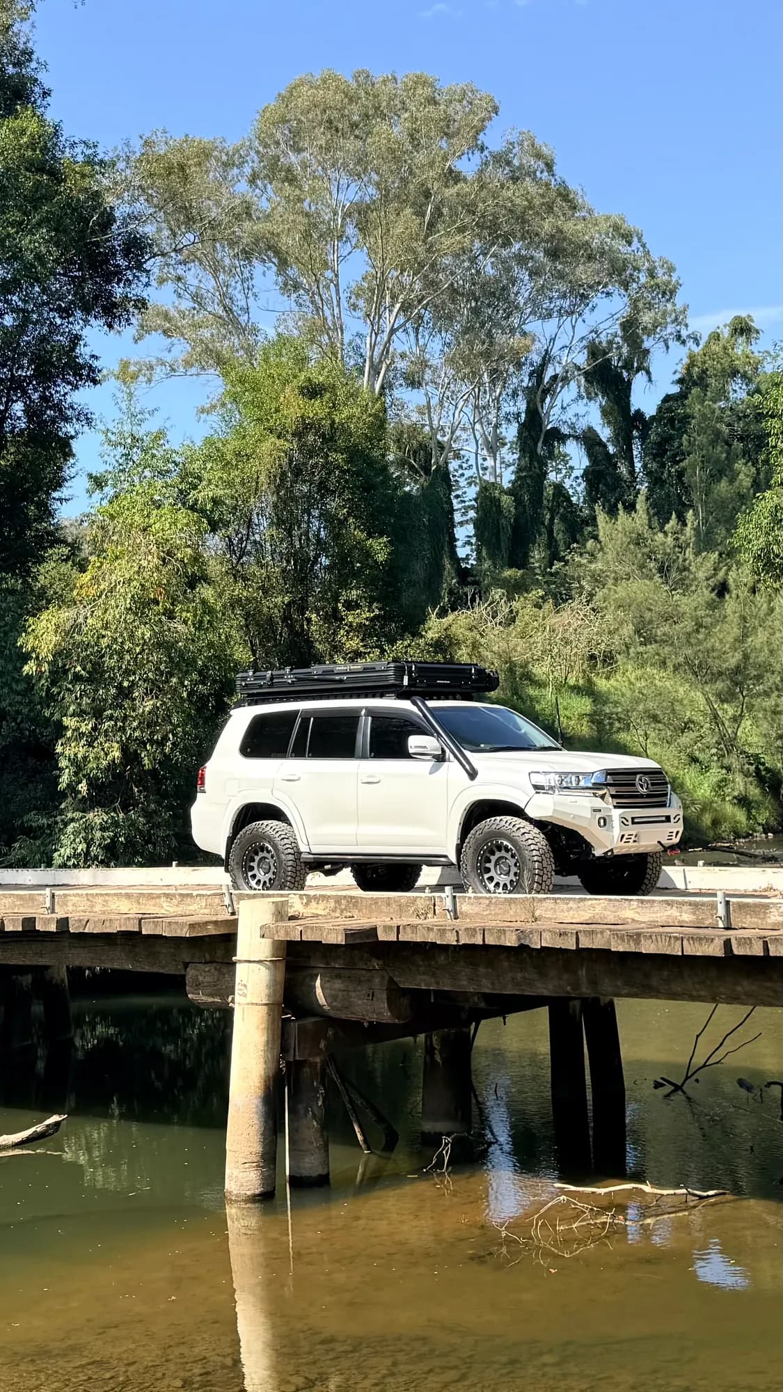 A white 200 Series with a rooftop tent crossing an old timber bridge over a creek