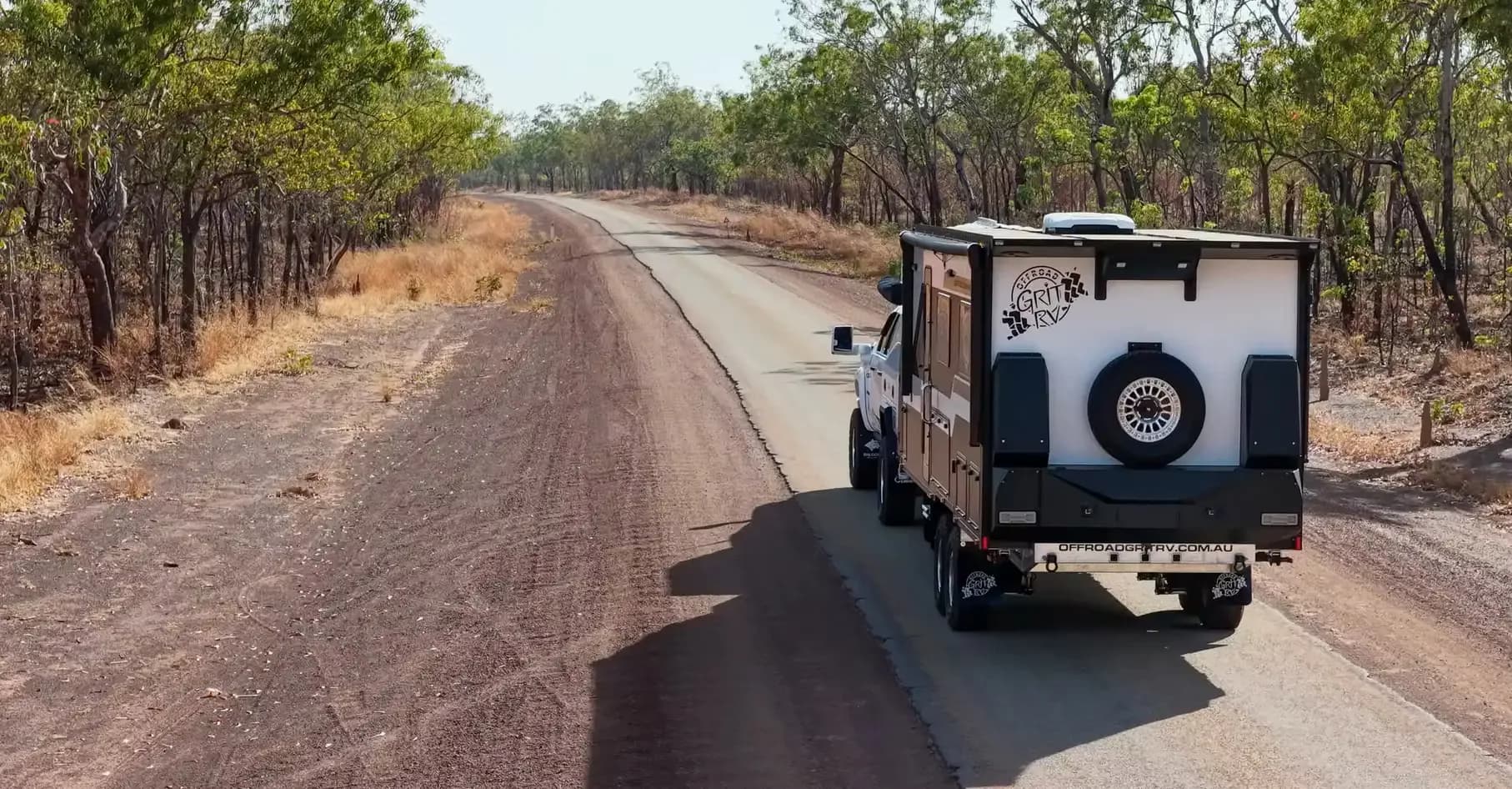 A 4x4 towing an off-road caravan along a gravel road through dry open woodland