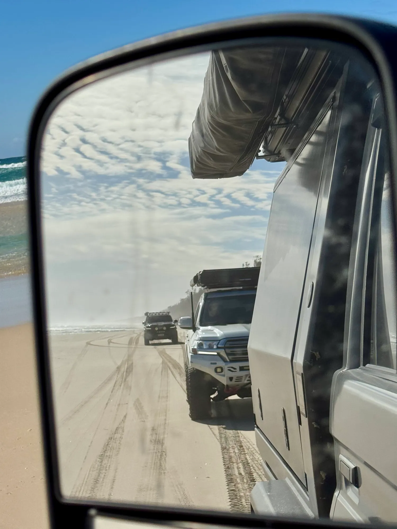 A side mirror showing a convoy of 4x4s following along a wide hard-sand beach