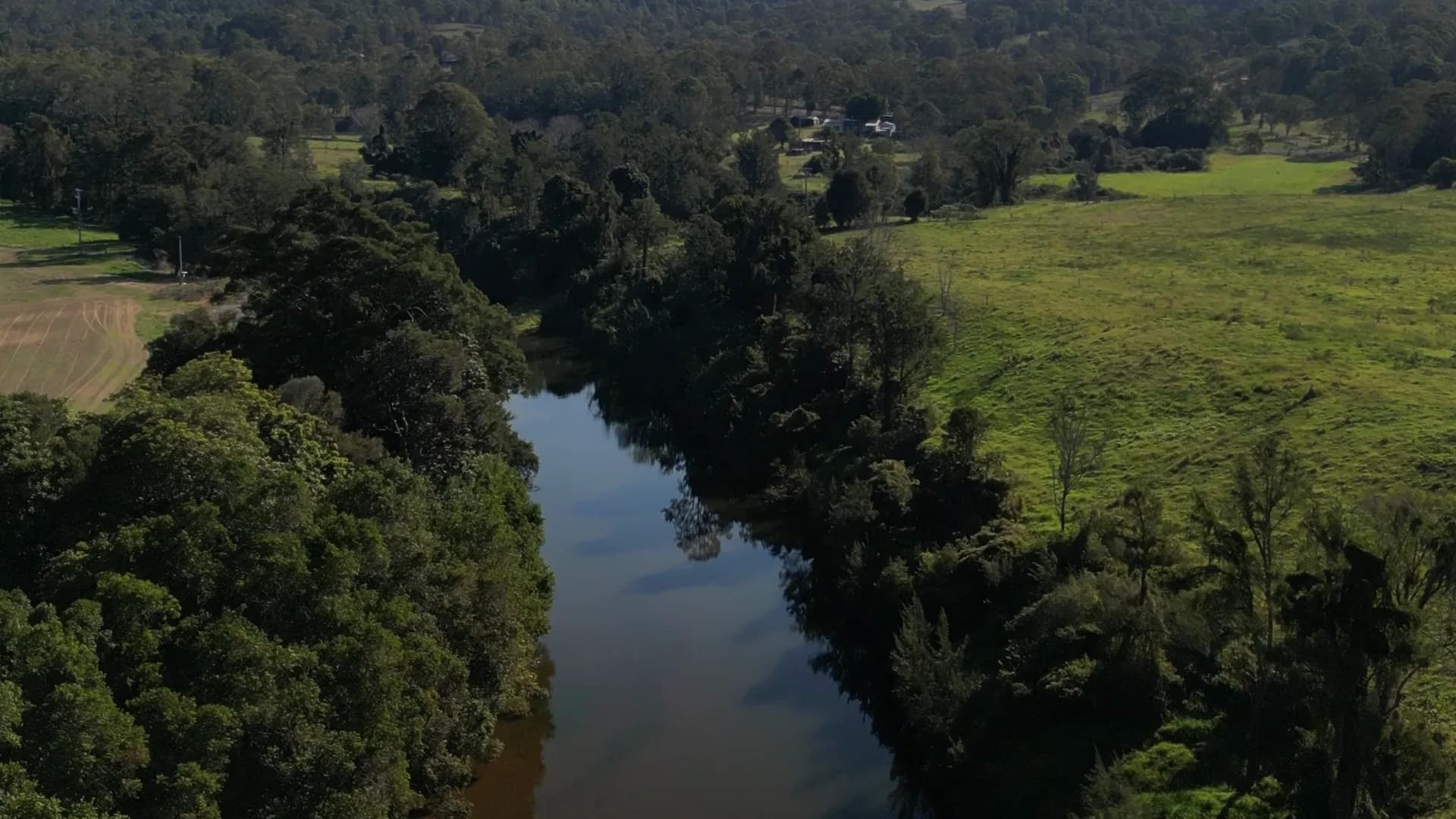 Drone view flying along a river through the trees, past green paddocks and homesteads