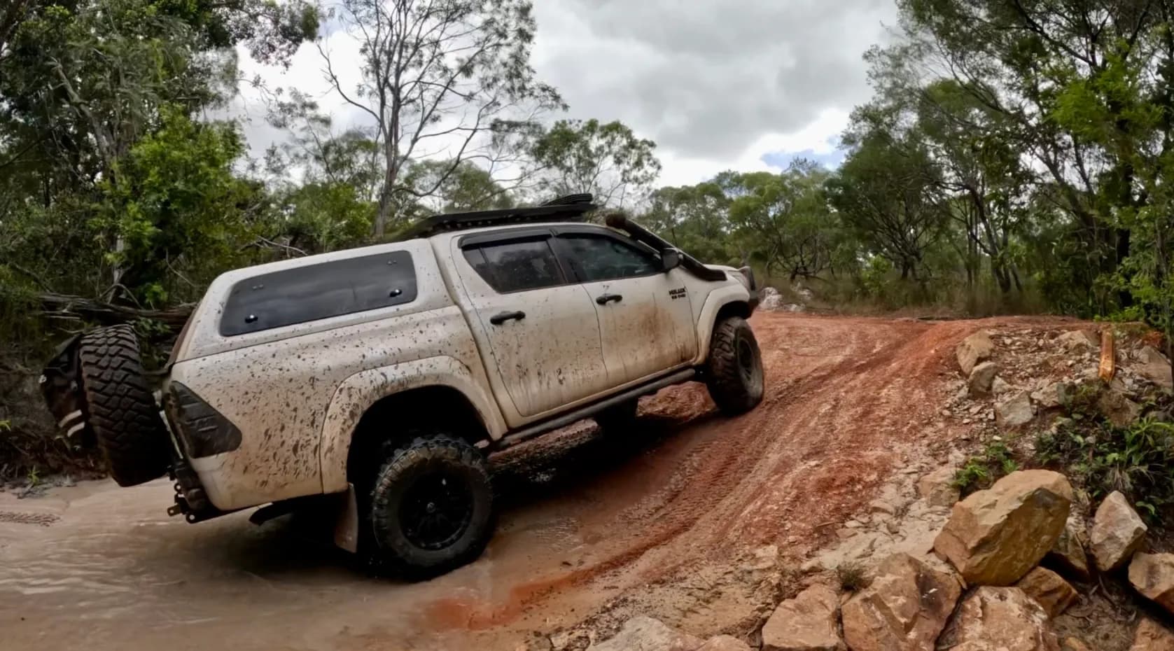 A mud-splattered white Hilux with a canopy climbing a rocky red dirt bank out of a shallow creek
