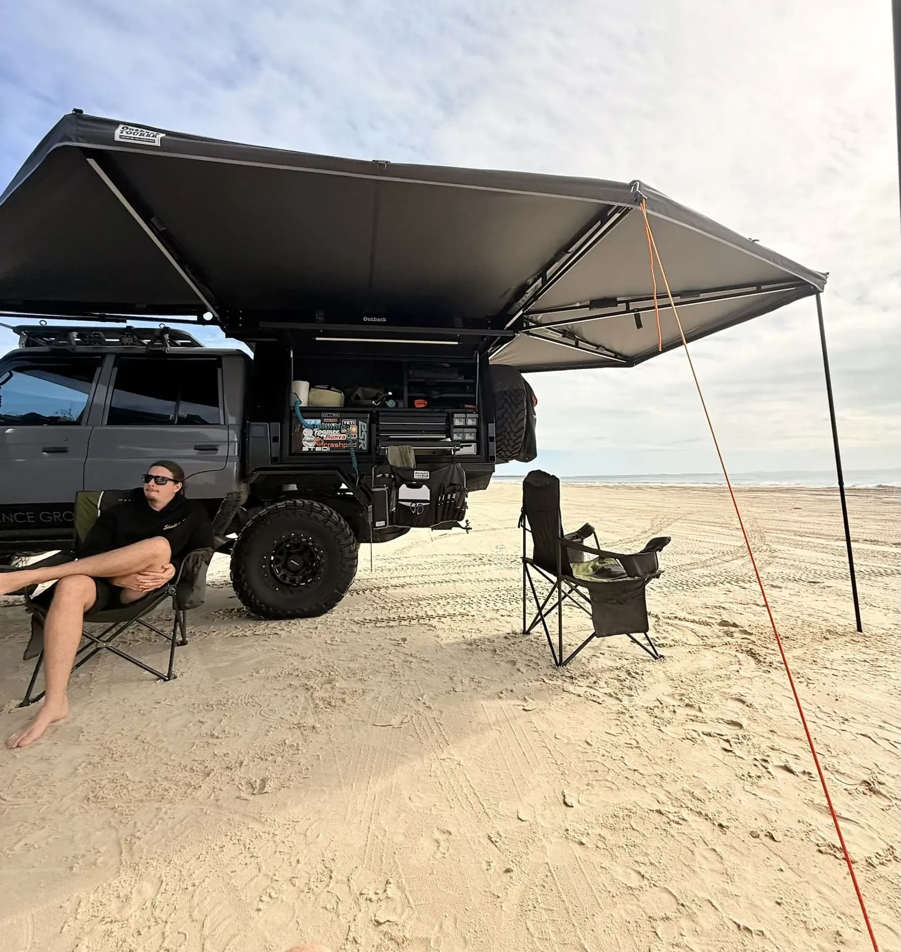 Sam relaxing in a camp chair under the awning of a dark 79 Series on a North Stradbroke Island beach